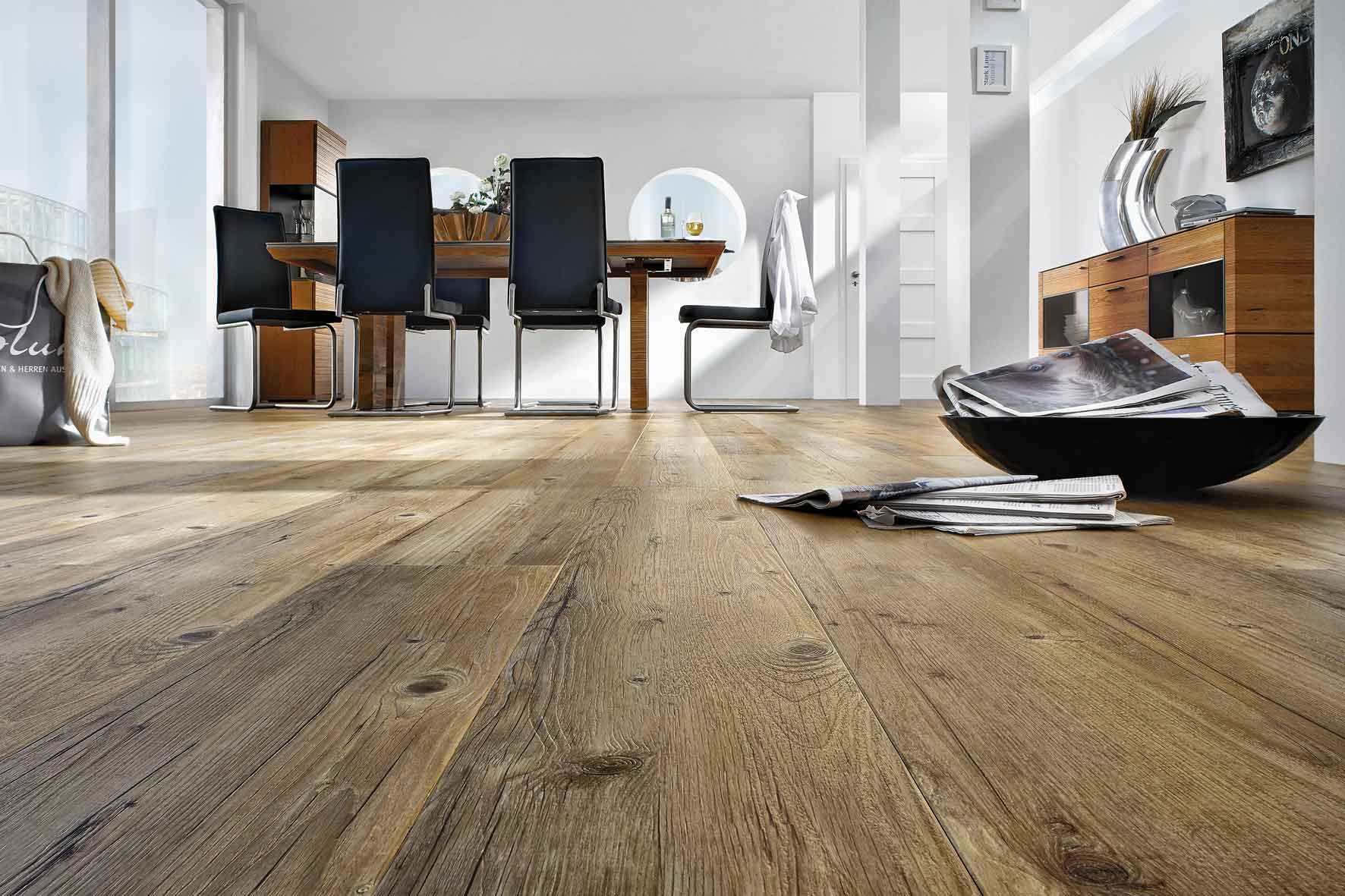Esszimmerbereich mit einem Tisch und sechs schwarzen Swingsesseln mit grauen Beinen auf braunem Holzoptikboden. Im Vordergrund steht eine Schüssel auf dem Boden, die mit Zeitungen gefüllt ist.