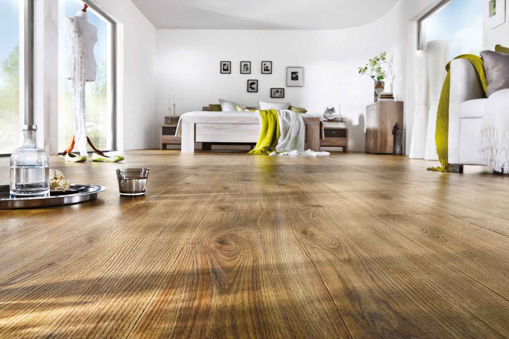 Schlafzimmer mit Holzoptikboden; im Vordergrund stehen ein Tablet, eine Wasserflasche und ein Glas direkt auf dem Boden. Im Hintergrund ein Bett mit grüner und weißer Bettwäsche, darüber sechs Bilder an der Wand. Links und rechts neben dem Bett jeweils ein Nachtschrank, daneben eine Kommode. Am rechten Bildrand ist eine Couch angeschnitten zu sehen.