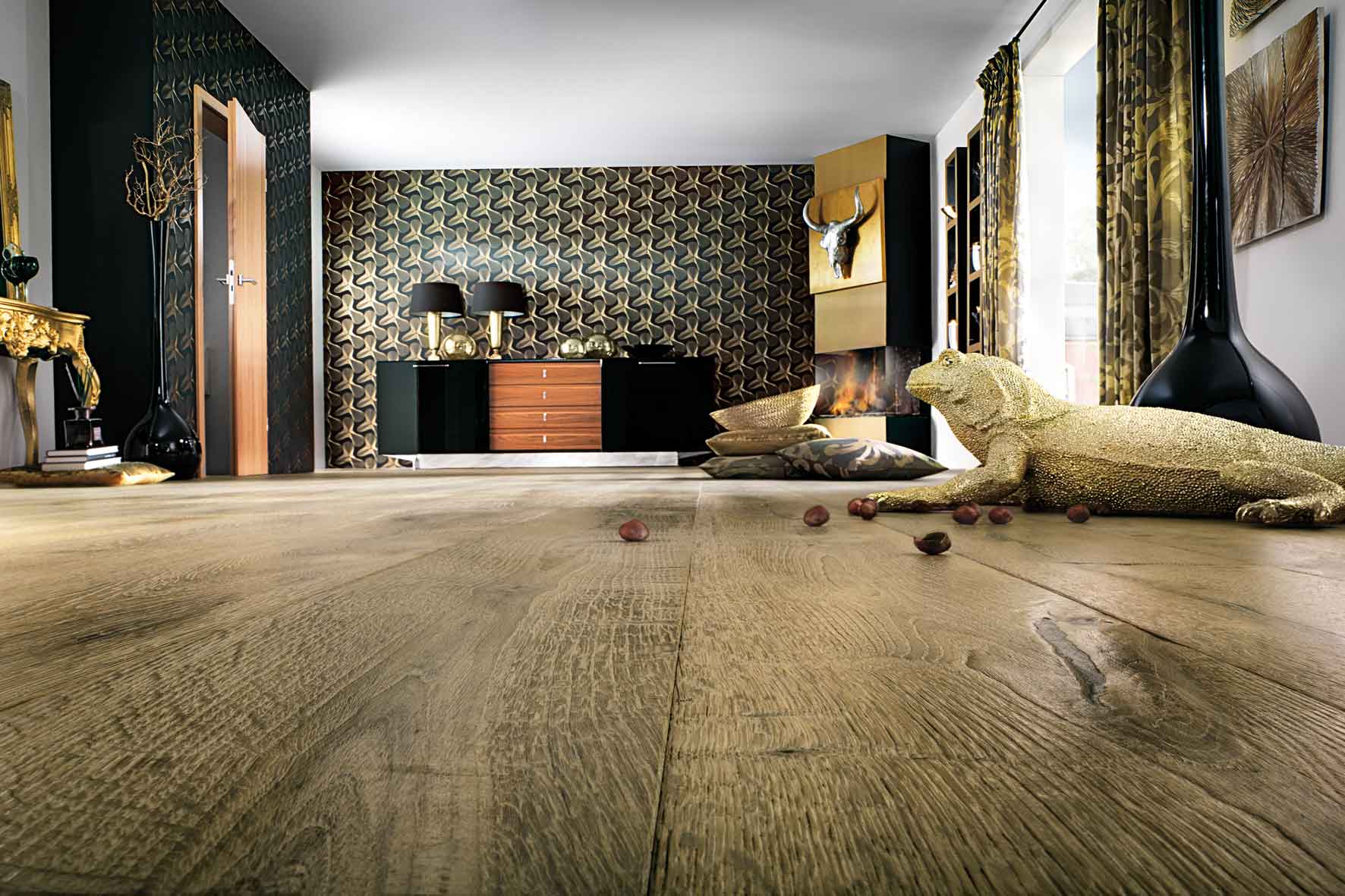 Raum mit Holzoptikboden und zwei tapezierten Wänden. An einer Wand steht eine Kommode, in der rechten Ecke befindet sich ein goldfarbener Kamin mit einem Tiergeweih darüber. Im Vordergrund liegen eine goldene Echse und eine schwarze Vase auf dem Boden.