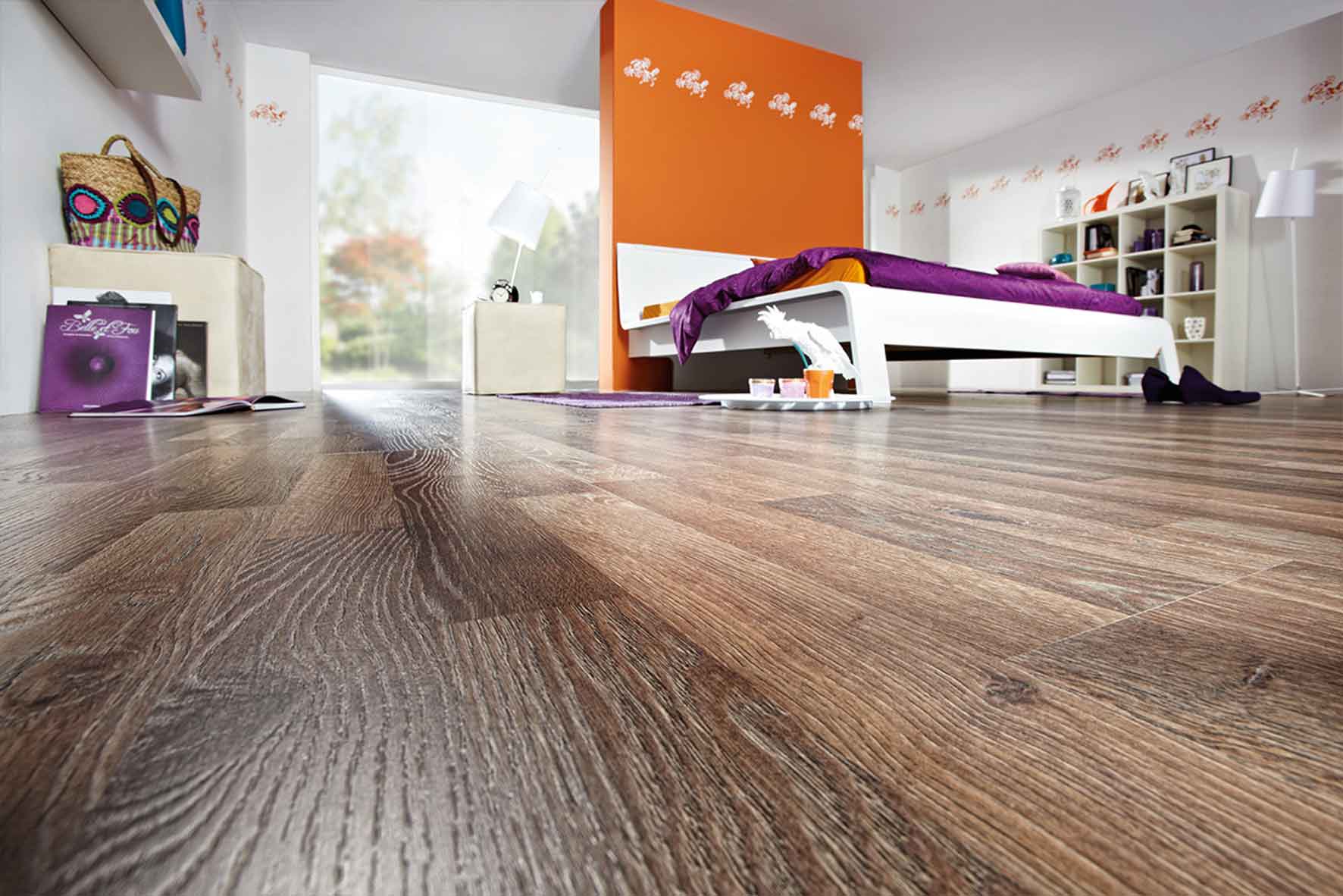 Schlafzimmer mit dunkelbraunem Laminatboden; im Raum steht eine orangefarbene Trennwand, davor ein weißes Bett mit pinkfarbener und oranger Bettwäsche.