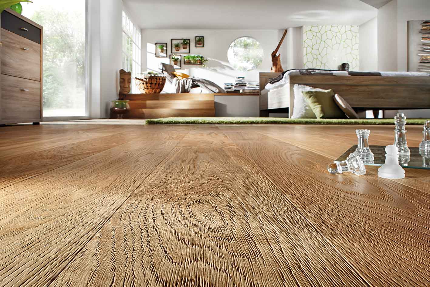 Schlafzimmer mit zwei Ebenen: Auf der unteren Ebene mit Holzbodenoptik stehen ein Holzbett und ein Glasschachbrett im Vordergrund. Drei Stufen führen hinauf zur oberen Ebene mit grünem Teppich, einem Beistelltisch, einer Liege und sieben an der Wand hängenden Pflanzen in schwarzen Rahmen.