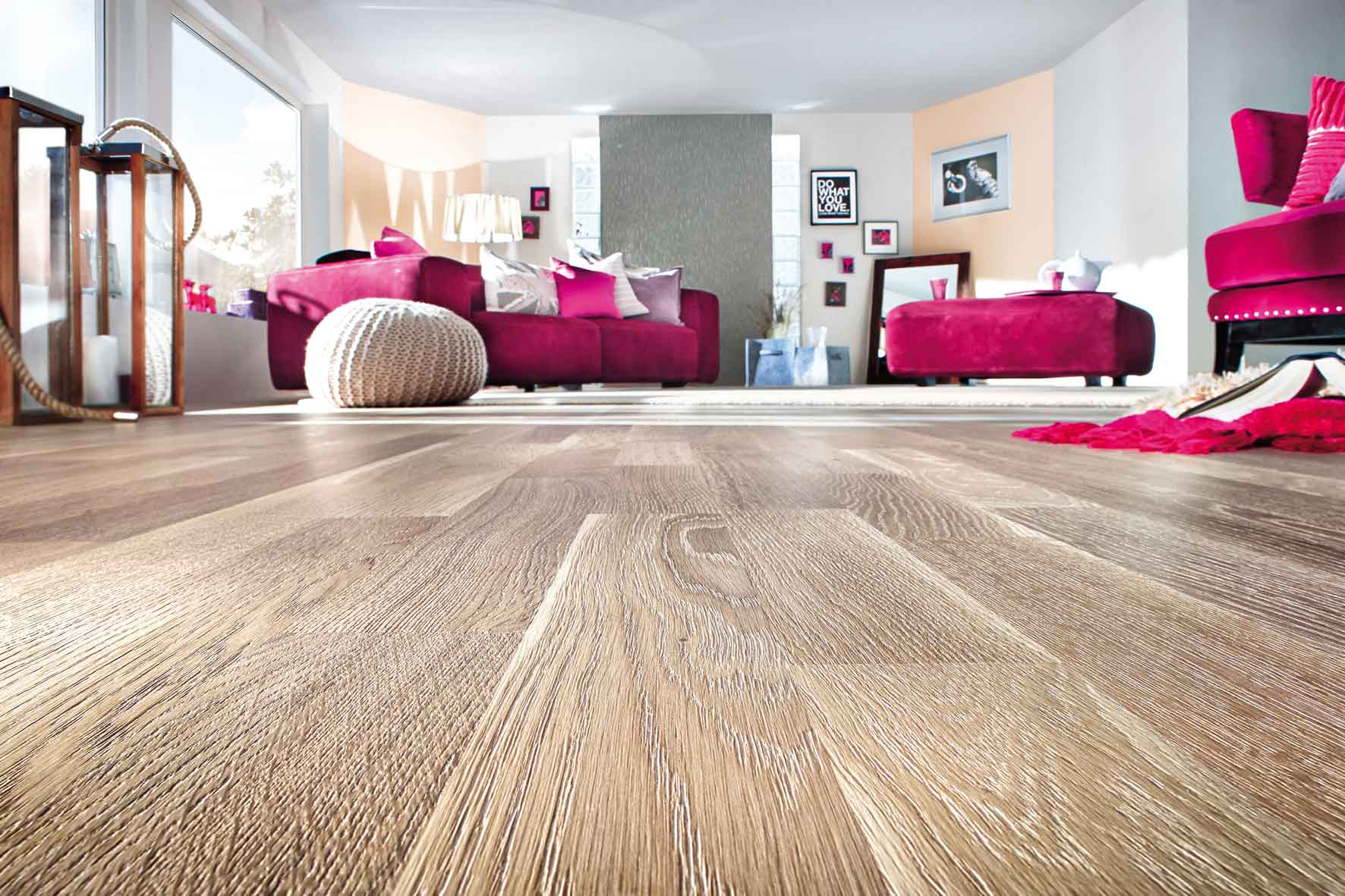 Wohnzimmer mit Holzoptikboden, pinkem Dreisitzer-Sofa, passendem Hocker und pinkem Sessel. Sofa und Hocker stehen auf einem beigen Teppich, davor befindet sich ein runder Geiger-Pouf als zusätzliche Sitzgelegenheit. Im Hintergrund hängen acht Bilder an der Wand.