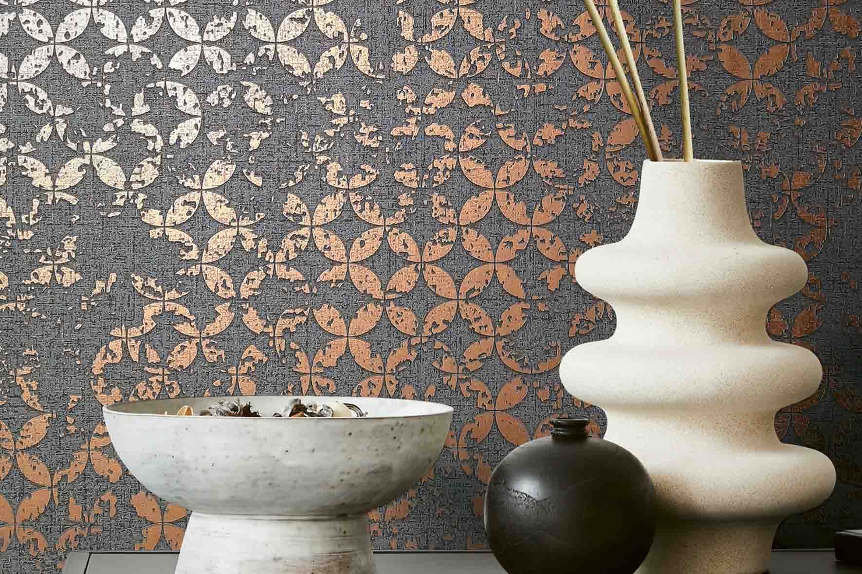 Dekorative Vase mit ungewöhnlicher Form neben einer Schale mit getrockneten Blättern vor einer Wand mit geometrischem Muster in grau und orange metallic.