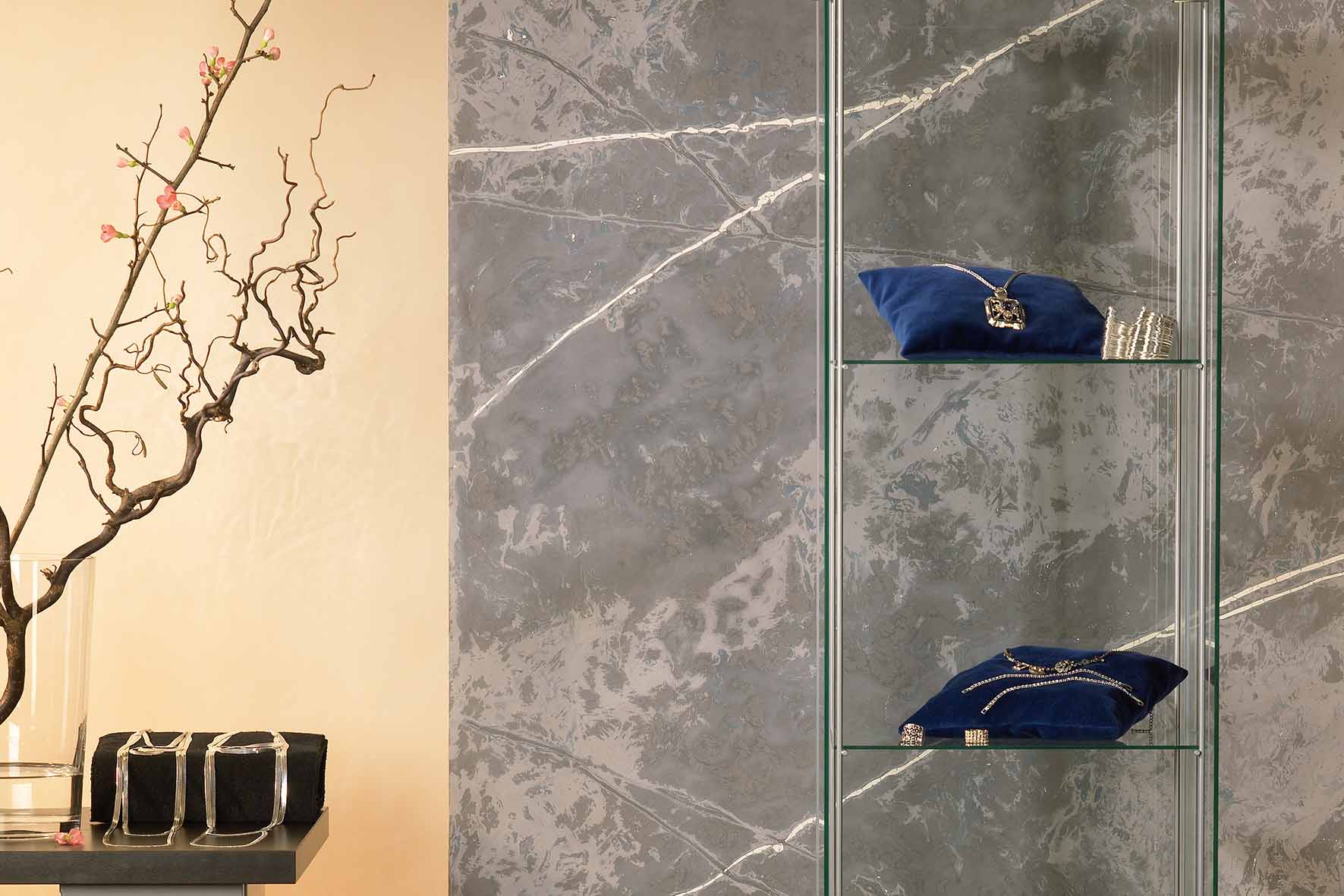 Vitrine mit zwei blauen Samtkissen, darauf Schmuckstücke, neben einem Tisch mit schwarzem Kissen mit silbernen Schmuck und Zweig mit Knospen, vor einer Wand in Betonoptik und orange.