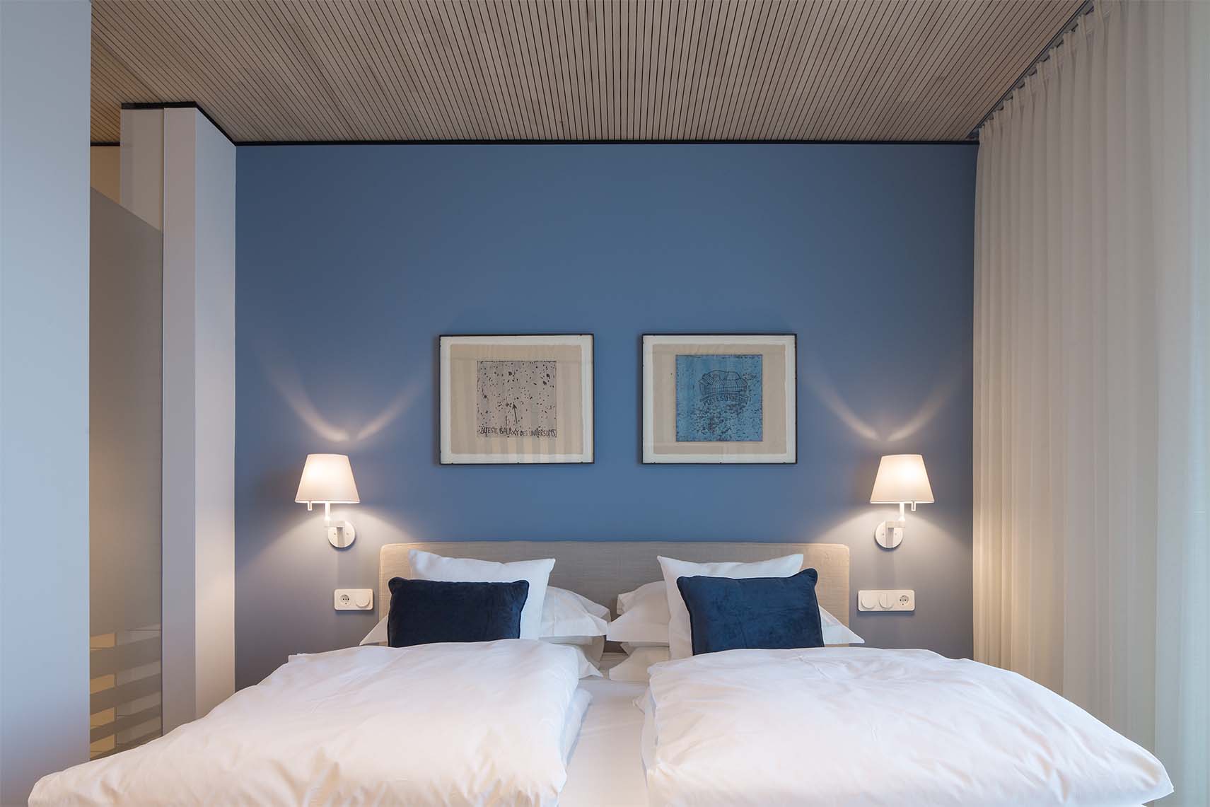 Doppelbett mit weißen Bettdecken und blauen Kissen vor einer blauen Wand mit zwei gerahmten Bildern und zwei Wandlampen.