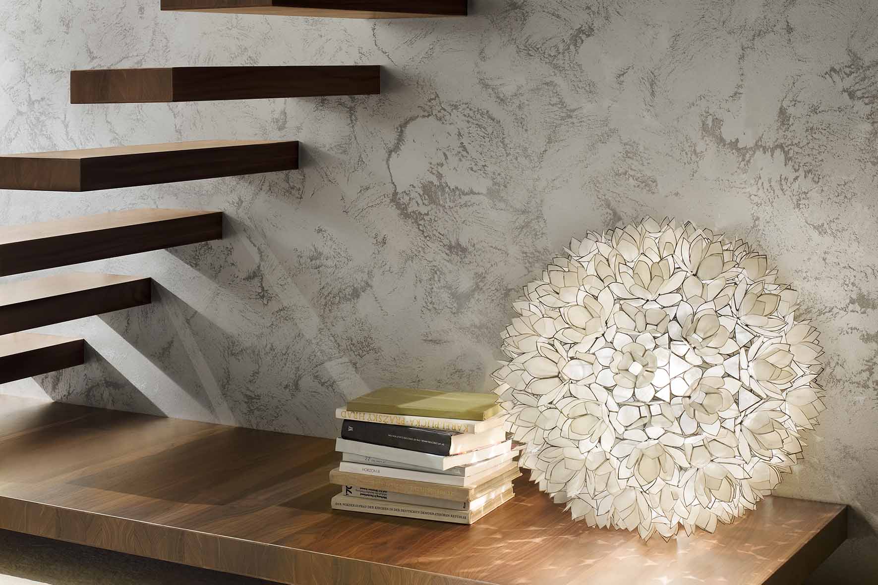 Schwebende Holztreppe über einem Holzregal mit einem Stapel Bücher und einer kugelförmigen Lampe aus weißen Blütenblättern.