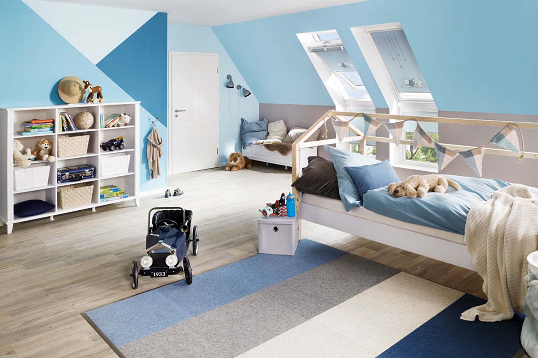 Kinderzimmer mit Dachschrägen, blau-weißer Wandgestaltung, Holzbett in Hausform mit blauer Bettwäsche und Stofftieren, Regal mit Spielzeug und Bücher, Spielauto auf dem Boden.