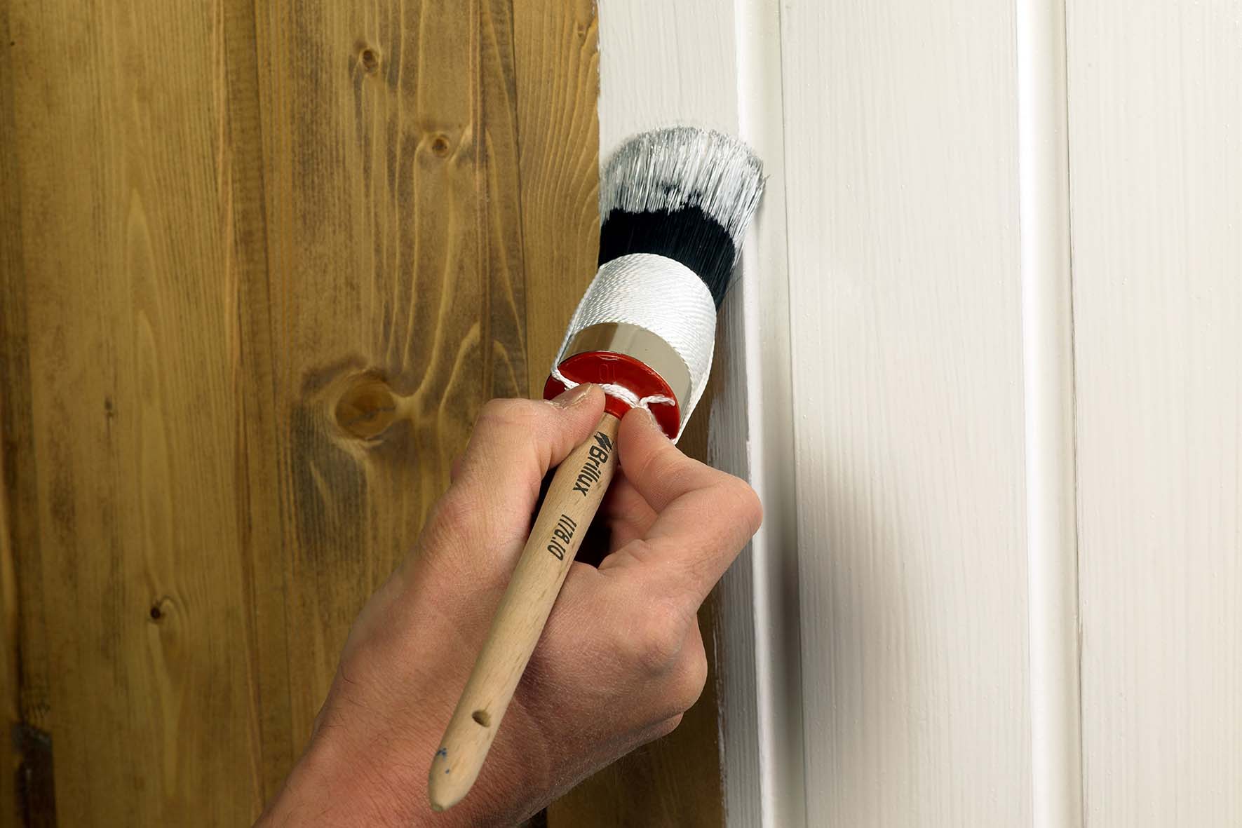 Hand streicht mit rundem Pinsel weiße Farbe auf eine Holzoberfläche neben unbehandeltem Holz.