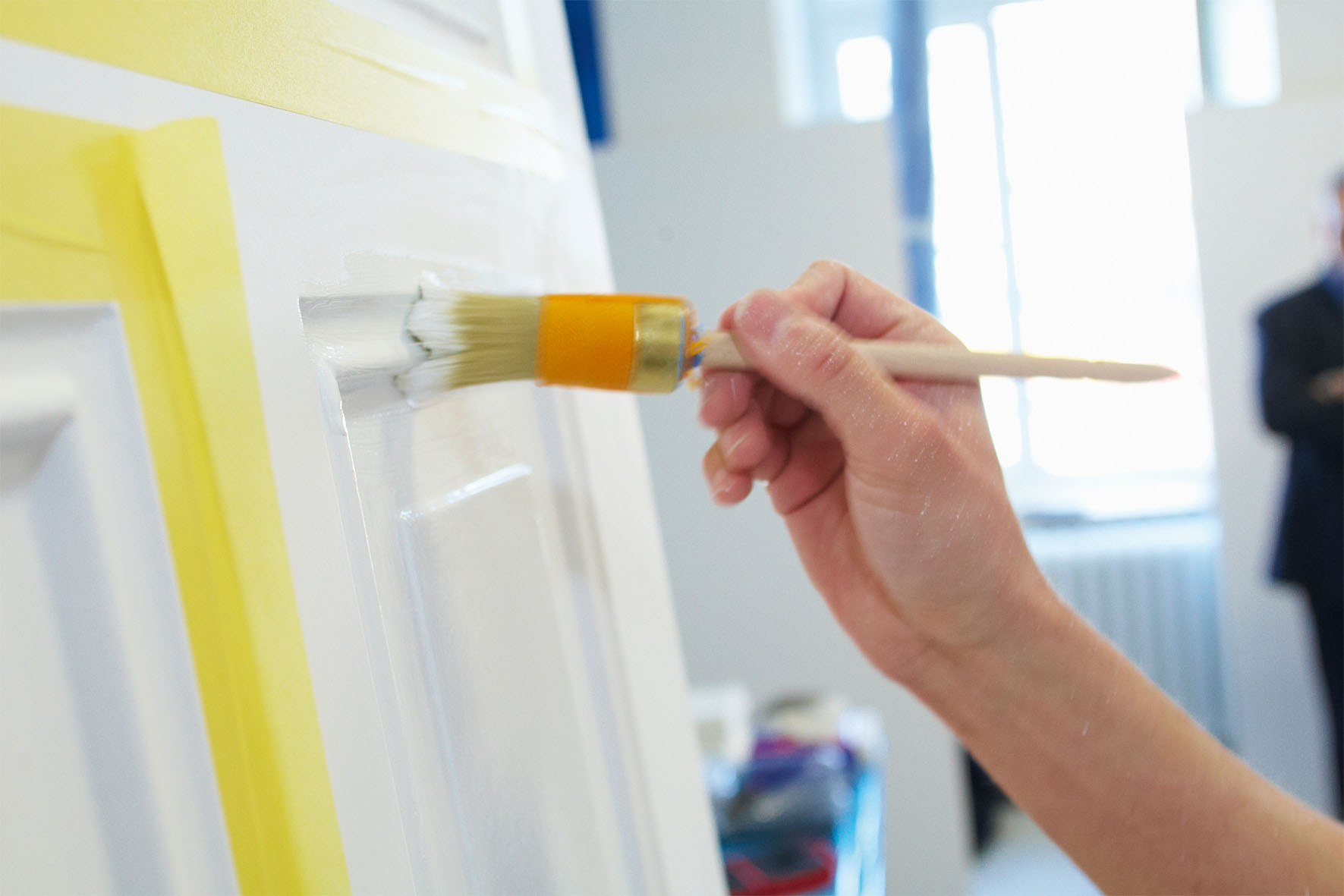Hand mit Pinsel streicht weiße Farbe auf eine Tür mit abgeklemmten Kanten.