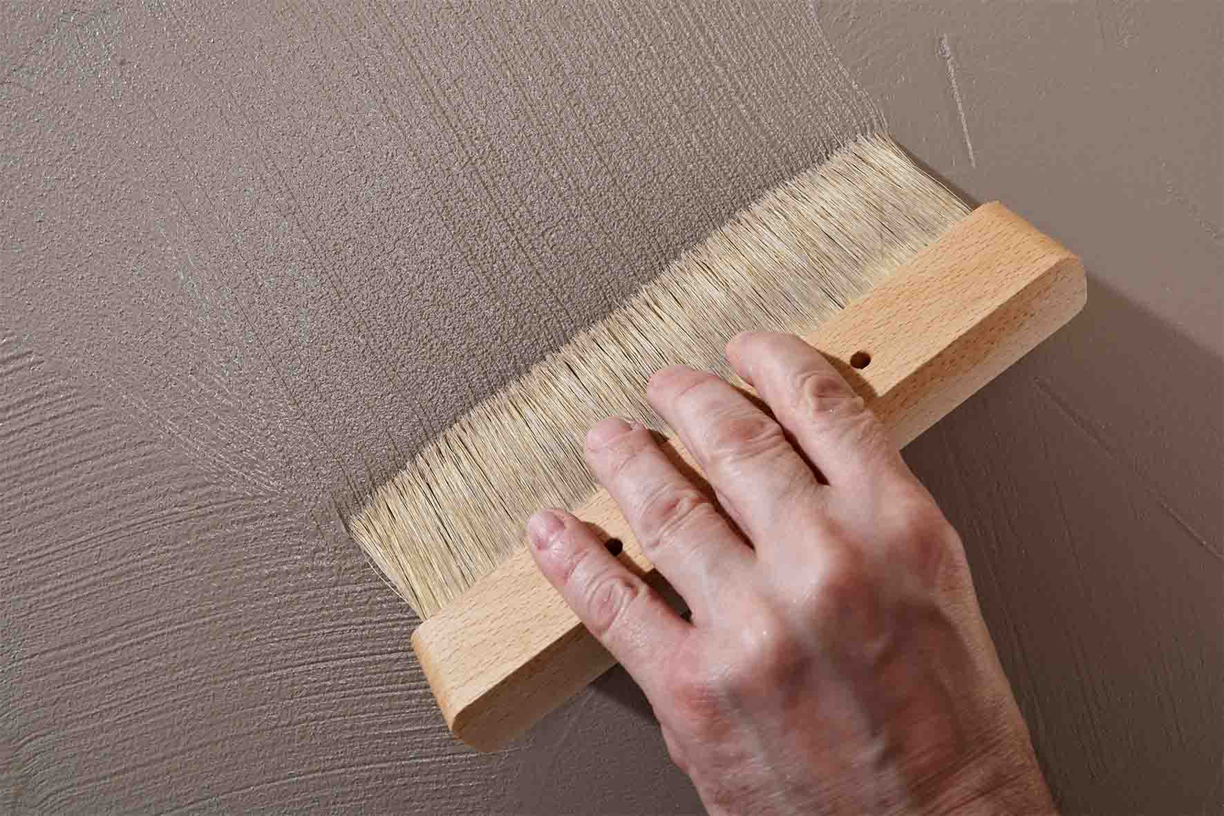 Hand hält breiten Malerpinsel und trägt braun graue Farbe auf Wand auf.