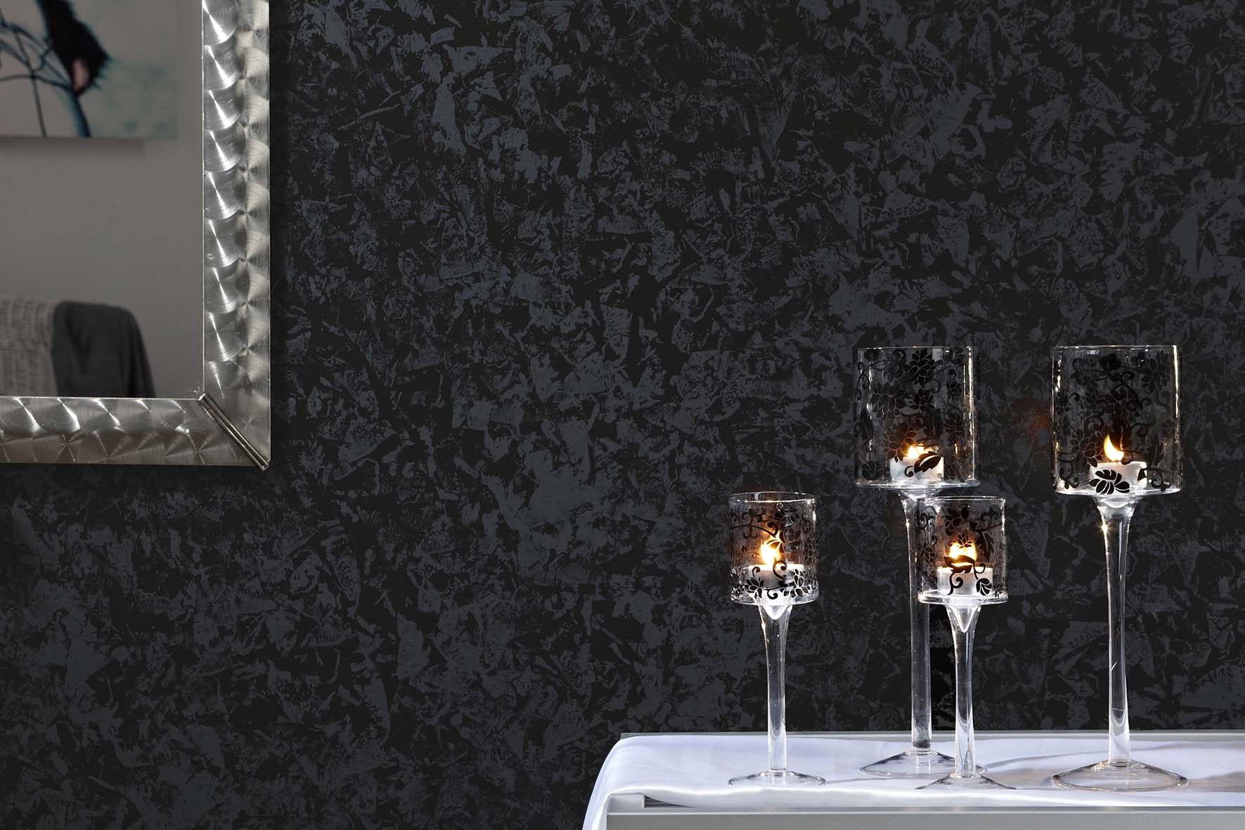Vier hohe Glaswindlichter mit brennenden Kerzen auf weißer Tischdecke vor schwarzer, strukturierter Wand.