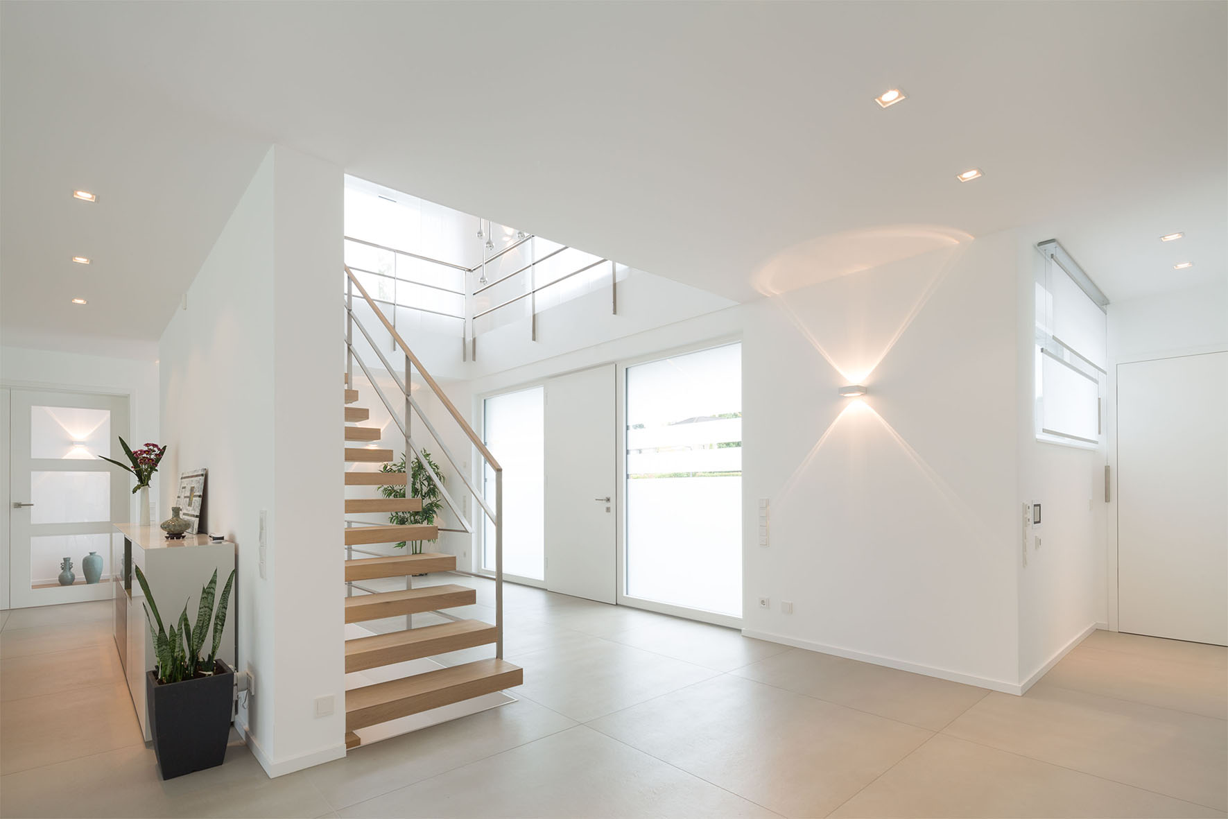 Heller Eingangsbereich mit Treppe aus Holz und Metallgeländer, großen Fenstern und weißen Wänden.