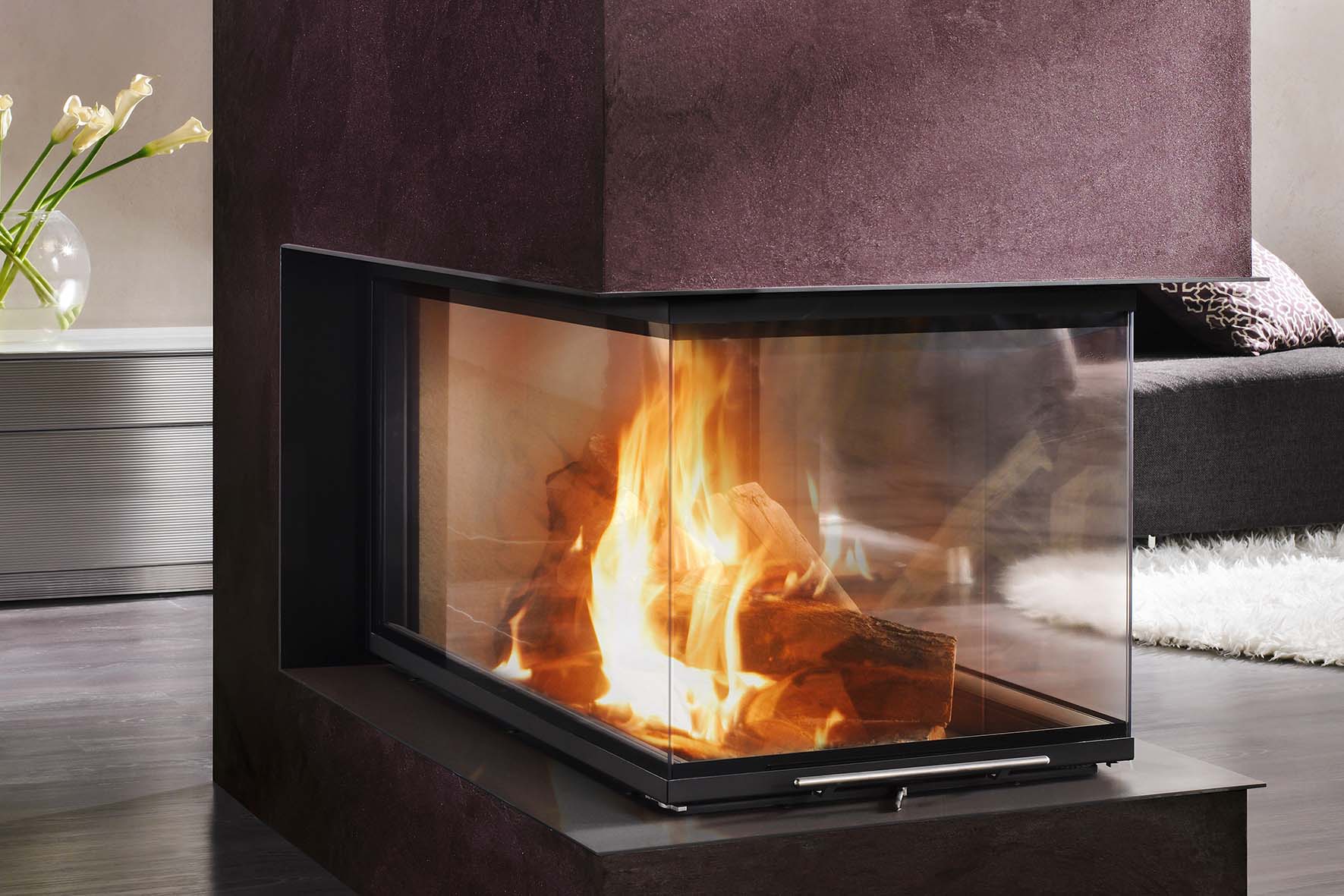 Eckkamin mit brennendem Holzfeuer in modernem Wohnzimmer mit dunklem Sofa und hellem Teppich.